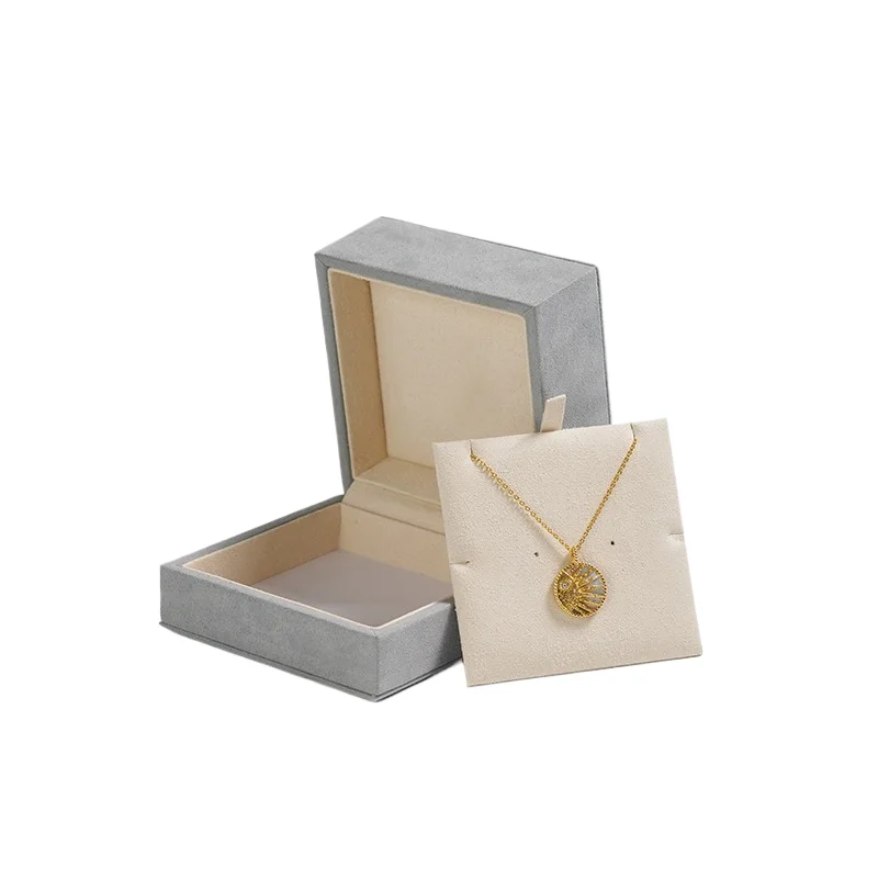 Grey suede flip-top pendant box opened back on its hinge beside the cream pendant display card holding a gold sunburst medallion necklace, showing the cream microfiber lining