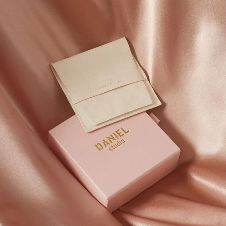 custom jewelry packaging — cream suede flap pouch beside a pink box with a gold foil logo