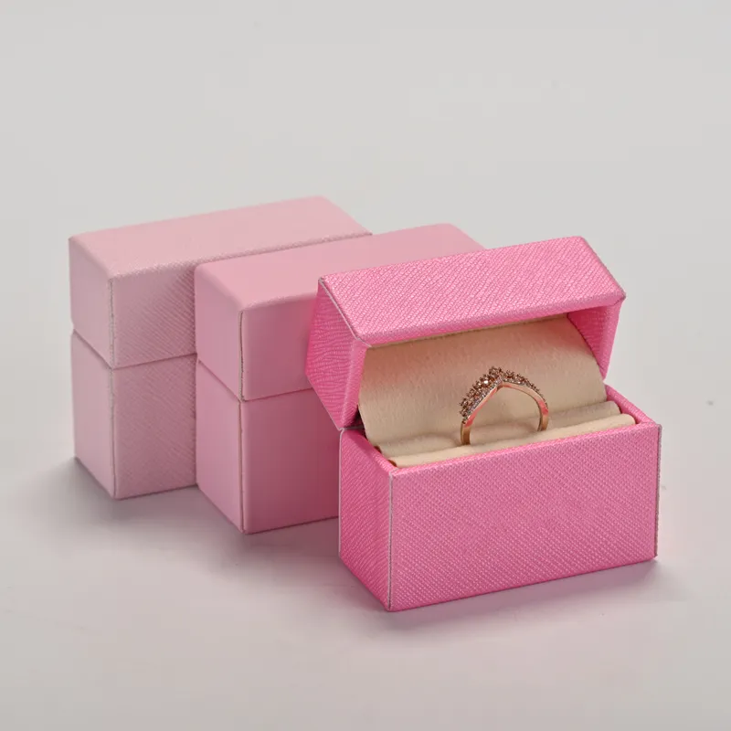 Three pink leather ring boxes in light pink, pink and hot pink saffiano texture, the front hot pink box open to reveal a cream velvet ring slot with a rose-gold pave wishbone ring