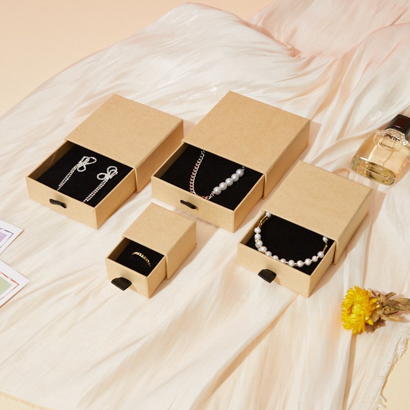 Cajas de joyería de cajón en papel kraft en varios tamaños con un collar, una pulsera y un anillo sobre almohadillas negras