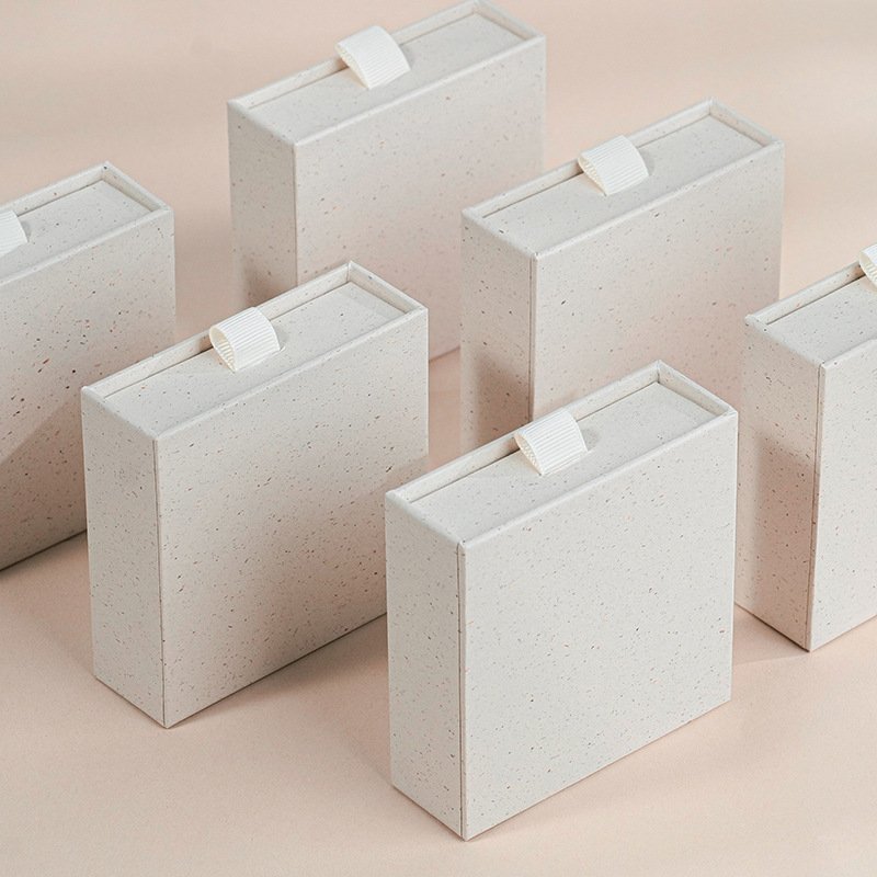 Cajas de joyería de cajón en papel blanco moteado con aspecto reciclado y lengüetas de listón, mostradas en grupo